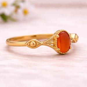 May include: A gold ring featuring an oval, orange gemstone. The ring has an ornate design with decorative elements on the band and around the gemstone. The ring is set against a soft, light background.