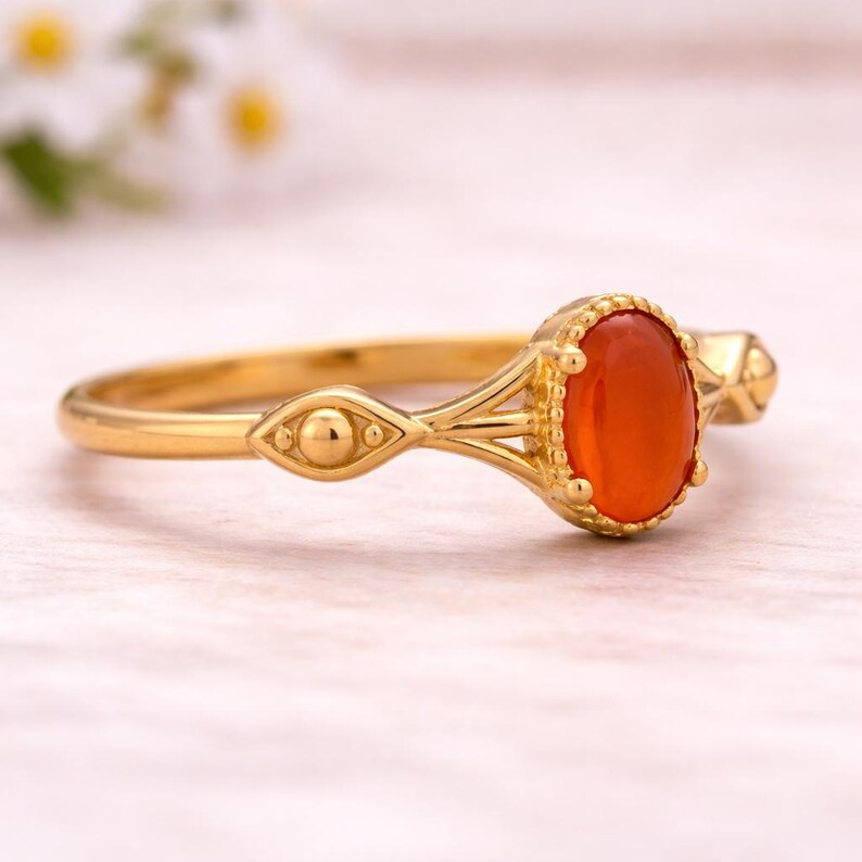 May include: A gold ring featuring an oval, orange gemstone. The ring has an ornate design with decorative elements on the band and around the gemstone. The ring is set against a soft, light background.