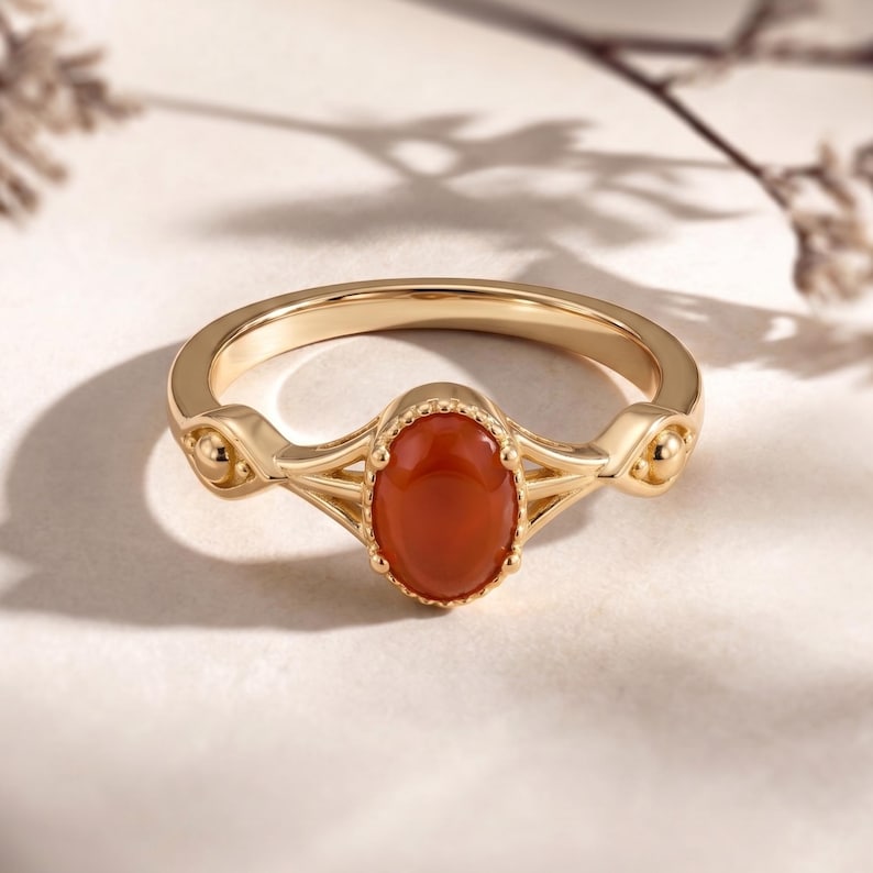 May include: A gold ring featuring an oval, reddish-orange gemstone. The ring has a delicate design with decorative elements on the band. The gemstone is set in a bezel setting, and the ring is displayed on a neutral background.