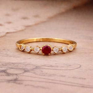 May include: A gold ring featuring a central round red gemstone, flanked by smaller white gemstones. The ring has a delicate band and is set against a neutral background, showcasing the jewelry's design and craftsmanship.