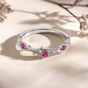 Op de afbeelding: Een zilveren ring met een doornontwerp, met drie ronde, helder roze edelstenen. De band van de ring heeft een textuur, verweven takkenpatroon. De edelstenen zijn gelijkmatig verdeeld.