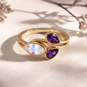 Op de afbeelding: Een gouden ring met een marquise-geslepen, iriserende maansteen en twee peervormige, dieppaarse amethist edelstenen. De ring heeft een vloeiend ontwerp, met de edelstenen in een cluster gezet. De ring wordt gepresenteerd op een lichtgekleurd oppervlak.