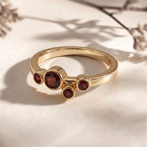 Könnte beinhalten: Ein goldener Ring mit fünf runden, tiefroten Edelsteinen. Der Ring hat ein glattes, poliertes Band und die Edelsteine sind in einzelnen Fassungen gefasst. Das Design ist elegant und modern, geeignet für den Alltag oder besondere Anlässe.