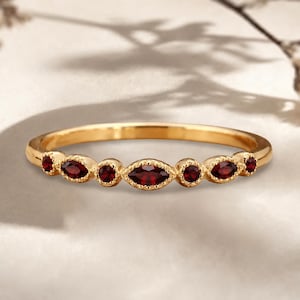 Könnte beinhalten: Ein goldener Ring mit einer Reihe von sieben abwechselnd marquise- und rundgeschliffenen, tiefroten Granat-Edelsteinen. Die Steine sind in eine zarte Metallfassung mit Milgrain-Rand eingefasst, die ein durchgehendes dekoratives Muster bildet.