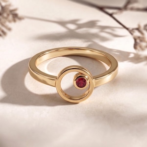 May include: A gold-colored ring with a circular design. The ring features a small, round, red gemstone set within a smaller circle. The band is smooth and polished, reflecting light. The ring's design is minimalist and modern.