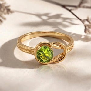 May include: A gold ring with a knot design, featuring a round, light green gemstone. The ring has a polished finish and a delicate band with subtle grooves. The gemstone is the focal point, set in a secure bezel.