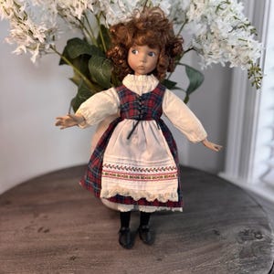 Puede incluir: Muñeca de porcelana con cabello rizado castaño y ojos azules, vestida con un vestido tradicional. El vestido presenta un corpiño a cuadros rojos y verdes, un delantal blanco con detalles bordados y zapatos negros. La muñeca posa con los brazos extendidos.