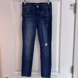 Puede incluir: Vaqueros de mezclilla azul oscuro con detalles desgastados, incluyendo roturas en las rodillas y áreas descoloridas. Los vaqueros tienen cierre de botón y cremallera, con bolsillos y trabillas. Los vaqueros están colgados en una percha plateada.