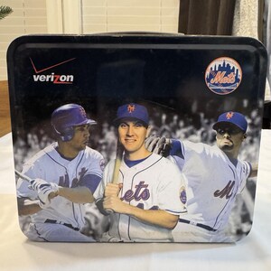 Puede incluir: Una lonchera rectangular de metal negro con el equipo de béisbol de los Mets de Nueva York. La lonchera tiene imágenes de jugadores de béisbol con sus uniformes. El logotipo de Verizon es visible en la esquina superior izquierda y el logotipo de los Mets en la superior derecha.