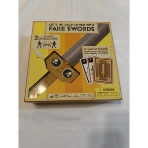 Peut inclure: Jeu de cartes en boîte intitulé "Let's Hit Each Other With Fake Swords". La boîte présente un dessin d'épée de dessin animé et comprend deux épées en mousse. Le jeu est pour 3-6 joueurs, à partir de 7 ans, et est un jeu de société familial.