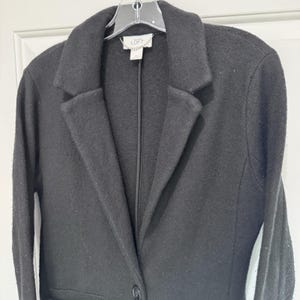 May include: A black blazer with a notched lapel and a single-button closure. The blazer is hanging on a clear hanger. The label reads "LOFT". The jacket has long sleeves and a tailored fit. The fabric appears to be a wool blend.