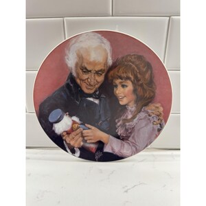 May include: A decorative plate featuring a vintage illustration of an older man and a young girl. The man, in a dark coat, is holding a toy, while the girl, in a pink dress, looks on. The background is a reddish-pink.