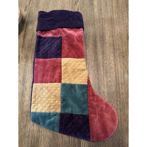 May include: A patchwork Christmas stocking in shades of burgundy, purple, gold, and teal. The stocking is made of velvet and features a quilted pattern on some of the squares. The cuff is a deep purple color.
