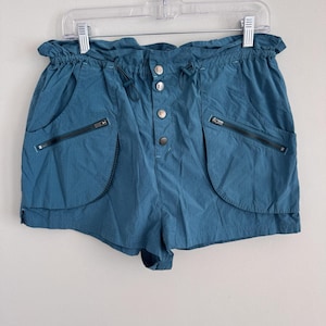 May include: Teal shorts with a drawstring waist and button fastening. The shorts have two front pockets with zip closures. The garment is suspended from a clear plastic hanger.