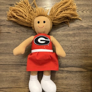 Puede incluir: Muñeca hecha a mano con piel bronceada, cabello rubio de hilo y una cara sonriente. La muñeca viste un atuendo de animadora rojo con una franja blanca y un logotipo "G" en blanco y negro. Los calcetines y zapatos blancos completan el look.