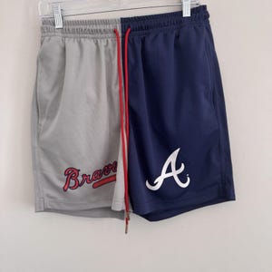 Puede incluir: Pantalones cortos deportivos con diseño dividido. Un lado es gris con "Braves" en rojo, el otro azul marino con un logo "A" blanco. Los pantalones cortos tienen un cordón rojo y están colgados de una percha.