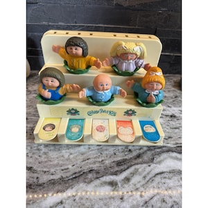 Op de afbeelding: Een vintage Cabbage Patch Kids muziekspeelgoed. Het speelgoed heeft zes Cabbage Patch Kids poppen op een gelaagde, crèmekleurige basis. Elke pop draagt een andere outfit. De basis heeft de woorden "Cabbage Patch Kids" en kleurrijke toetsen.