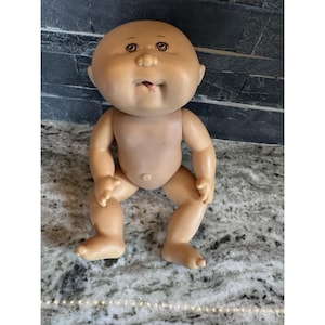 Peut inclure: Une poupée Cabbage Patch Kid vintage avec une peau bronzée, des yeux bruns et une petite bouche ouverte. La poupée est assise, les bras et les jambes tendus. Le corps de la poupée est fait d'une matière souple.