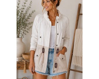 May include: A two-tone jacket with a white upper section and a tan lower section. The jacket has a stand-up collar, snap closures, and drawstring details. It features two front pockets and is worn with denim shorts.