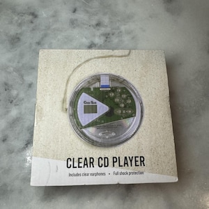 Può includere: Un lettore CD portatile trasparente, che mostra i componenti interni. Il lettore è nella sua confezione originale, che presenta la scritta "CLEAR CD PLAYER" e include auricolari trasparenti e protezione completa dagli urti.
