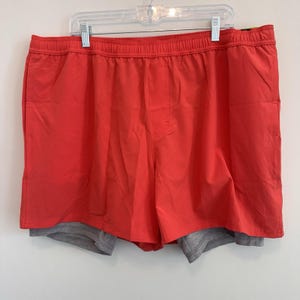 Puede incluir: Pantalones cortos deportivos de color rojo brillante con una capa inferior gris, colgados de una percha blanca. Los pantalones cortos tienen una cintura elástica y un pequeño logotipo en la pierna derecha. Los pantalones cortos están hechos de un material ligero.