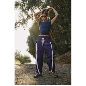 May include: A navy blue crop top and purple track pants with white side stripes. The pants have a drawstring waist. The person is wearing sunglasses and brown trainers. The background is a natural outdoor setting with trees and sunlight.