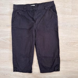May include: Black capri pants with a button closure at the hem. The pants have a button and zipper closure at the waist, with pockets on the front and back. The pants are made of a sturdy fabric.