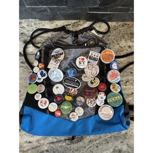 May include: A gray and blue drawstring backpack covered in various colorful pinback buttons. The buttons feature text and graphics, including "I Love NY," "Michigan Rocks," and "Jesus Texas & Tacos."