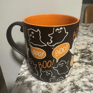 Op de afbeelding: Een zwarte keramische mok met een donkergrijze handgreep en een oranje binnenkant. De mok is versierd met witte cartoongeesten en oranje tekstballonnen met de tekst "BOO!". Een mok met Halloween-thema.