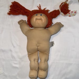May include: A vintage Cabbage Patch Kid doll with red yarn hair styled in pigtails. The doll has a tan skin tone, blue eyes, and a soft body. The doll's arms are outstretched, and it has a tag attached to its body.