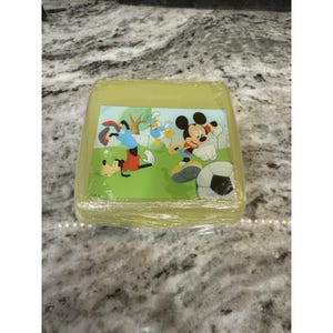 May include: A yellow plastic lunchbox featuring Mickey Mouse, Goofy, and Donald Duck playing soccer. The lunchbox is wrapped in clear plastic and has a green background with a soccer ball.