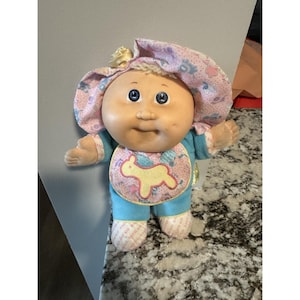 Peut inclure: Une poupée Cabbage Patch Kids vintage avec un visage en vinyle doux et des yeux bleus. La poupée porte une tenue bleue et rose, un bavoir avec un agneau jaune et un bonnet assorti. Les pieds de la poupée portent l'inscription "Cabbage Patch Kids".