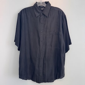 Puede incluir: Camisa negra de manga corta con cuello y bolsillo en el pecho. La camisa está colgada sobre un fondo blanco. La camisa tiene botones y mangas cortas.