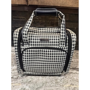 Puede incluir: Un bolso tote con estampado de cuadros blancos y negros con asa y ribete de cuero negro. El bolso tiene un bolsillo frontal con cremallera y una pequeña etiqueta rectangular. El bolso está sobre una superficie clara.