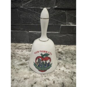May include: White ceramic bell with a long handle and gold trim. The bell features a colorful graphic of an apple with the New York City skyline and the words "NEW YORK CITY" and "THE BIG APPLE".