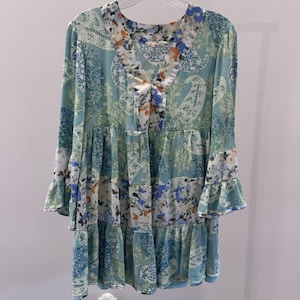 Puede incluir: Una blusa tipo túnica azul claro con cuello en V y mangas largas. La blusa presenta un diseño de patchwork con estampados florales y de cachemira en tonos azules, blancos y naranjas. Las mangas tienen un detalle de volantes en los puños.