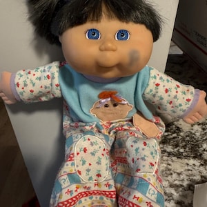 May include: A vintage doll with black pigtails, blue eyes, and fair skin. The doll is dressed in a light blue top with a felt applique and floral patterned pyjamas. The pyjamas have red, white, and blue accents.