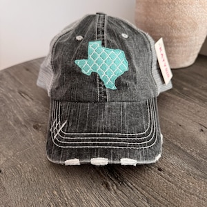 May include: A distressed black and gray baseball cap with a turquoise Texas-shaped patch featuring a white patterned design. The cap has a mesh back and white stitching details. A tag with the brand name "KATYBUG" is attached.
