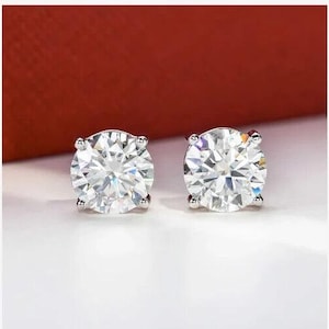 Puede incluir: Un par de pendientes de botón redondos de piedras preciosas transparentes. Las piedras preciosas están engastadas en un metal de color plateado. Los pendientes se exhiben sobre una superficie blanca con un fondo rojo.