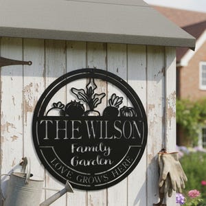 Könnte beinhalten: Schwarzes Metallschild für den Familiengarten mit dem Namen "The Wilson" und dem Text "Love Grows Here". Das Schild zeigt Gemüseillustrationen und ist an einem verwitterten weißen Holzschuppen befestigt. Eine Gießkanne und Gartengeräte sind in der Nähe.