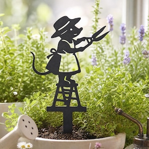 Könnte beinhalten: Ein schwarzer Gartenstecker aus Metall mit der Silhouette einer Maus, die einen Hut und eine Schürze trägt, auf einer kleinen Leiter steht und eine Gartenschere hält. Die dekorative Figur ist zum Einstecken in die Erde konzipiert.