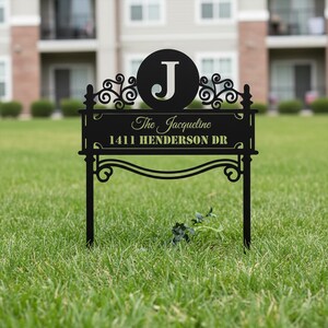 Könnte beinhalten: Schwarzes Metall-Gartenschild mit dem Namen "The Jacqueline" und der Adresse "1411 Henderson DR". Das Schild hat einen großen Buchstaben "J" in einem Kreis und dekorative Schnörkel. Es steht in einem Rasen.