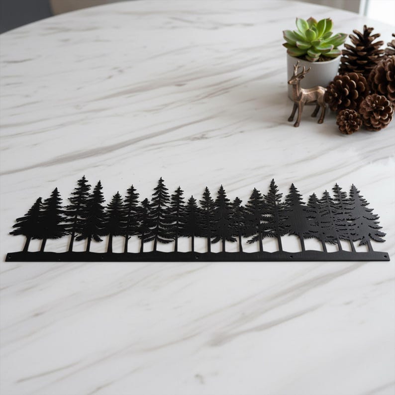 May include: Black metal forest silhouette. The decorative piece features a detailed cutout of a forest scene with various sizes of pine trees. The artwork is placed on a white marble surface, with a small succulent, deer figurine, and pine cones in the background.