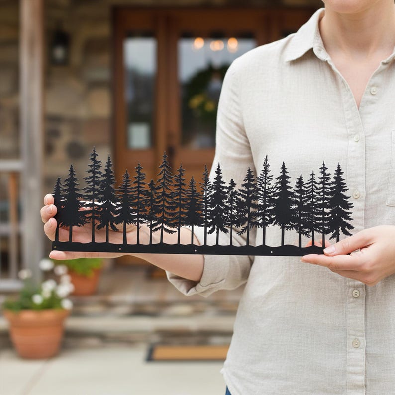 May include: Black metal wall art depicting a forest of pine trees. The silhouette design features detailed tree shapes and a horizontal base. This decorative piece is held in front of a house.