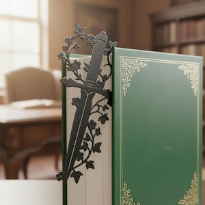 Könnte beinhalten: Ein schwarzer Metall-Lesezeichen in Form eines Schwertes mit floralen Akzenten, das in ein grünes Buch eingelegt ist. Das Lesezeichen hat eine Schwertklinge und Efeu. Das Buch hat goldfarbene, verzierte Eckendesigns.