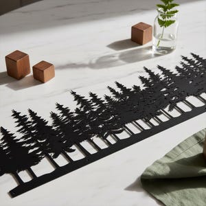 May include: A black metal table runner featuring a forest silhouette design. The runner is cut out to depict a line of evergreen trees. Two small wooden cubes and a small glass vase with a fern are also present.