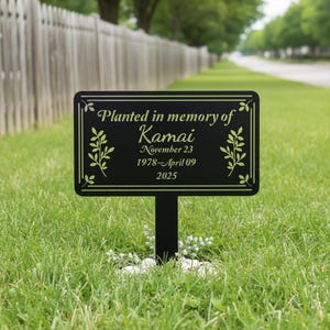 Peut inclure: Plaque commémorative noire plantée dans l'herbe, avec le texte "Planted in memory of Kamai" et des dates. La plaque a une bordure décorative et des motifs floraux. La plaque est montée sur un piquet noir.