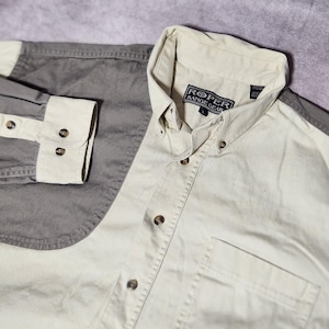 May include: A two-tone button-down shirt. The shirt has a light beige body and sleeves, with grey accents on the shoulders and cuffs. The cuffs have three brown buttons. The shirt has a chest pocket and the brand "ROPER RANGE GEAR" is visible on the collar.
