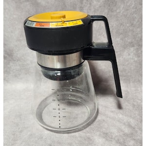 May include: A vintage coffee maker with a clear glass carafe and a black and yellow top. The top has a dial with settings for dark, medium, and light. The carafe has measurement markings for up to 12 cups.