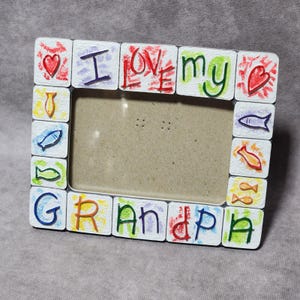 May include: A rectangular picture frame with a white textured border featuring hand-painted colorful designs. The frame displays the text I LOVE MY GRANDPA in multi-colored letters, interspersed with small painted red hearts and various fish.